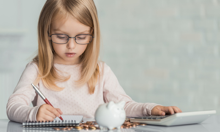 Little girl wearing eyeglasses writes in notebook at table witj calculator , coins and piggy bank
