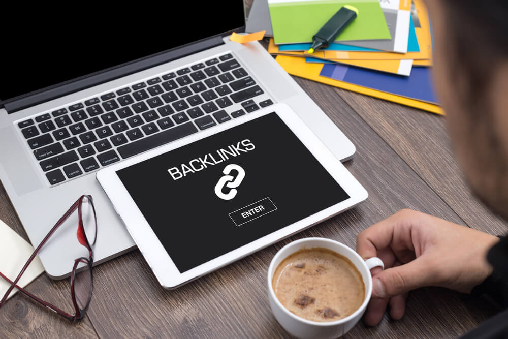 homem segurando xicara de cafe em frente a laptop e tablet com a palavra backlinks em tela