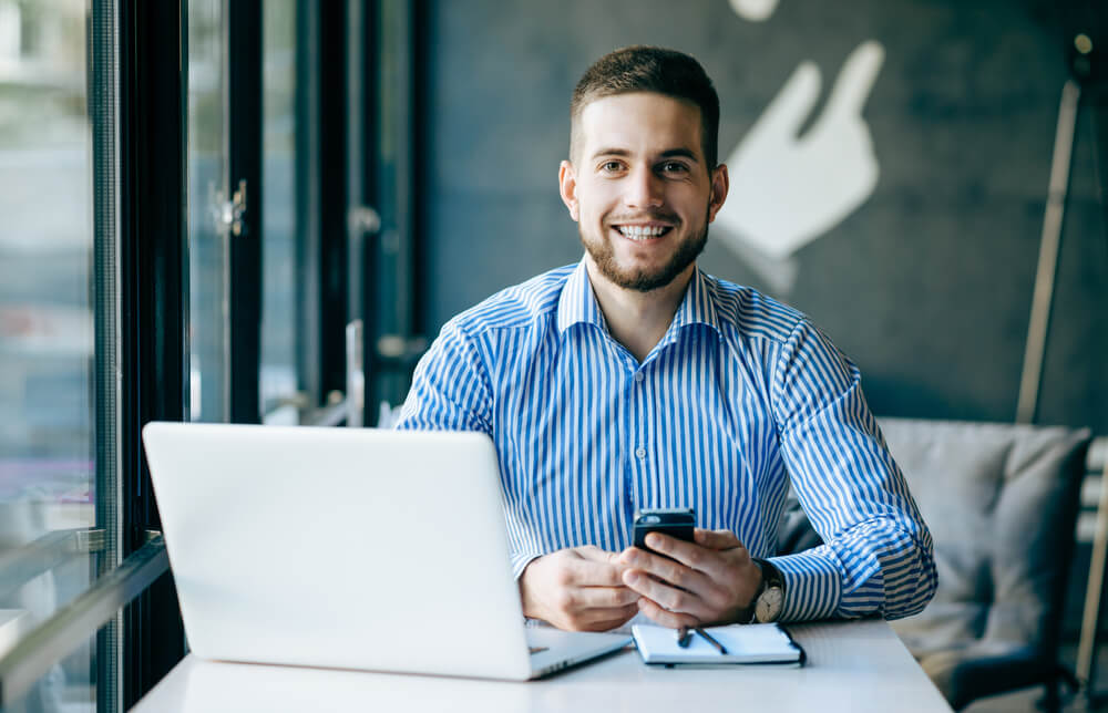 profissional sorridente em frente de laptop e usando smartphone