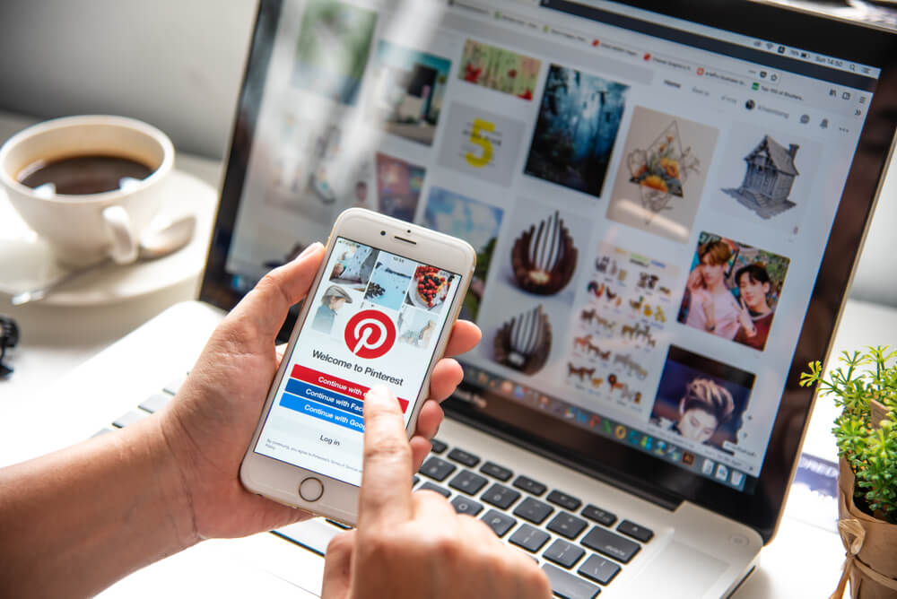 mao masculina segurando smartphone em frente a laptop com aplicativo pinterest em tela